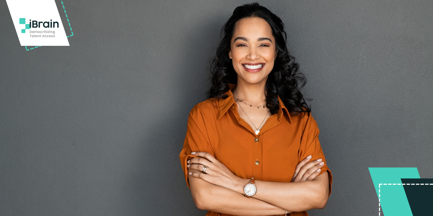 Woman of color smiling, wearing an orange short-sleeved dress shirt and dark gray wall in the background.